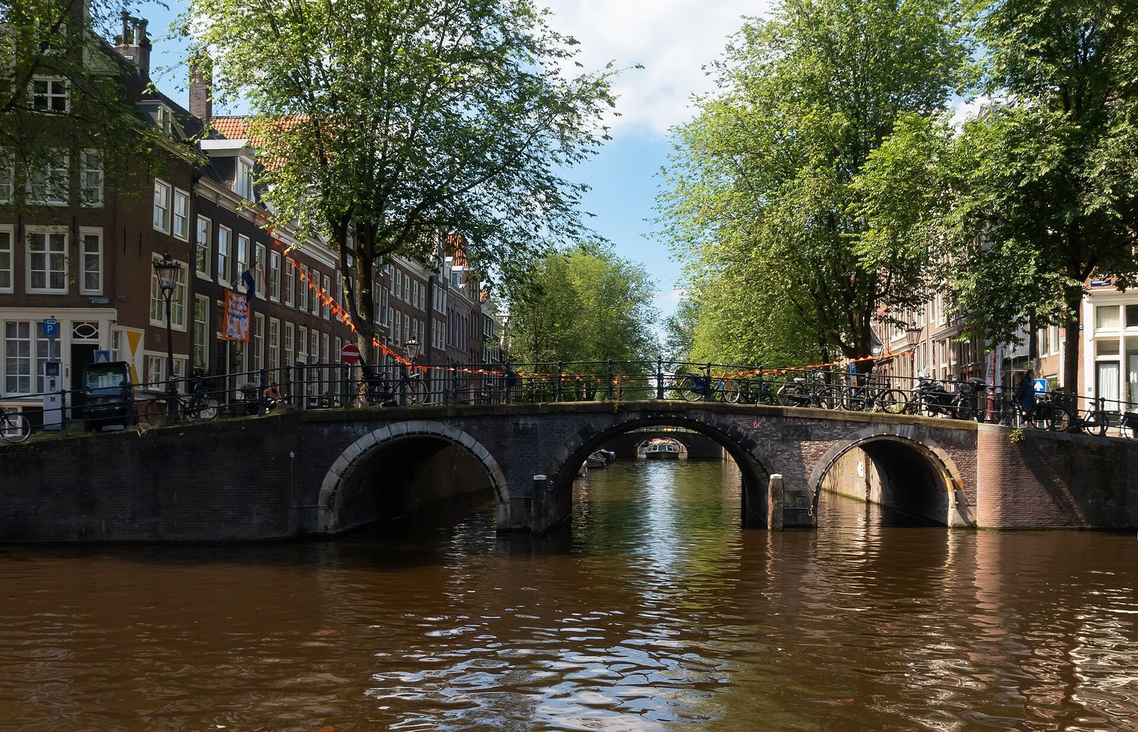 Eine dreibogige Backsteinbrücke über eine Gracht, davor orange Wimpel, links Grachtenhäuser, rechts Bäume