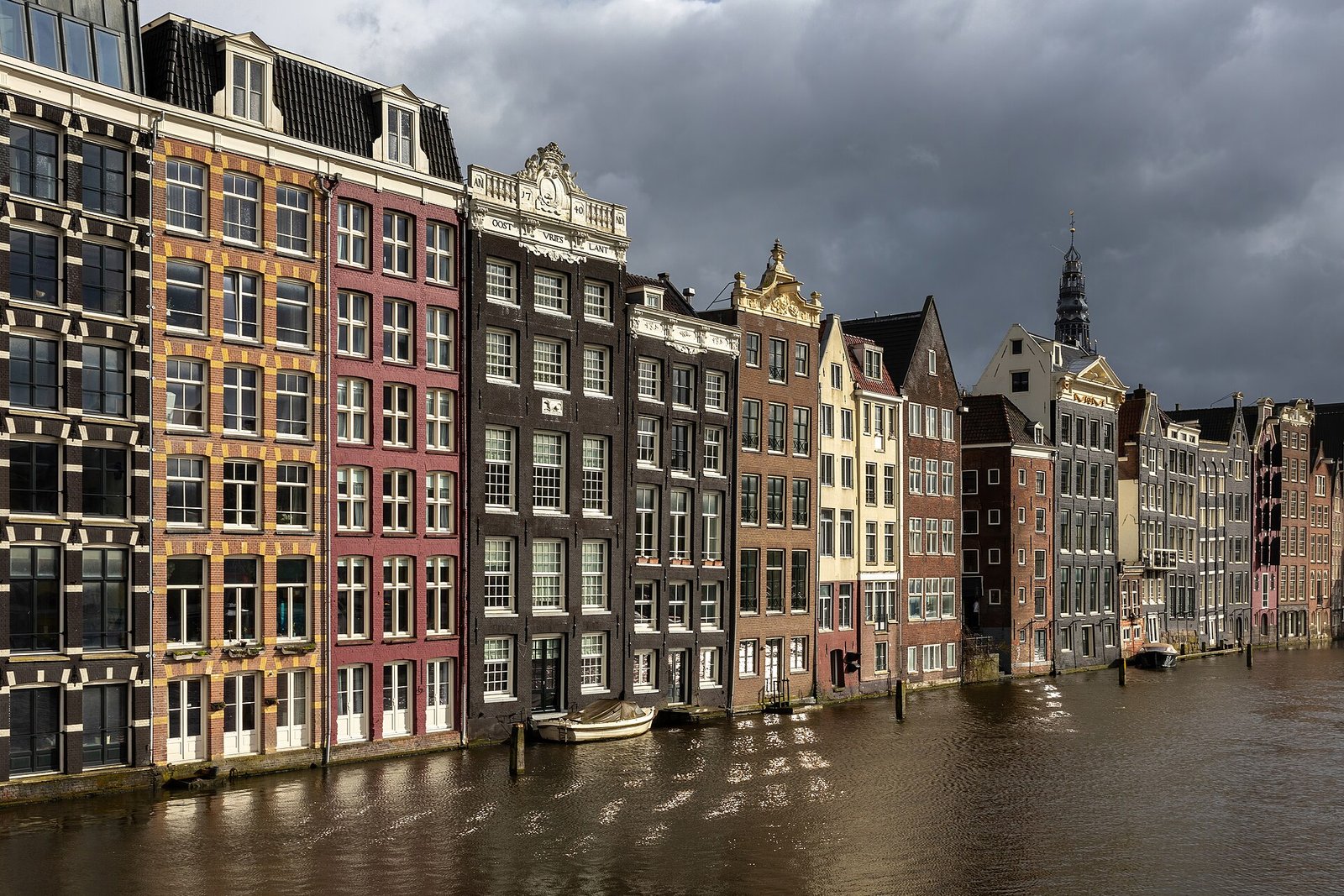 Eine Reihe schmaler Grachtenhäuser mit verzierten Giebeln am Wasser unter dunklen Wolken, rechts ein Kirchturm