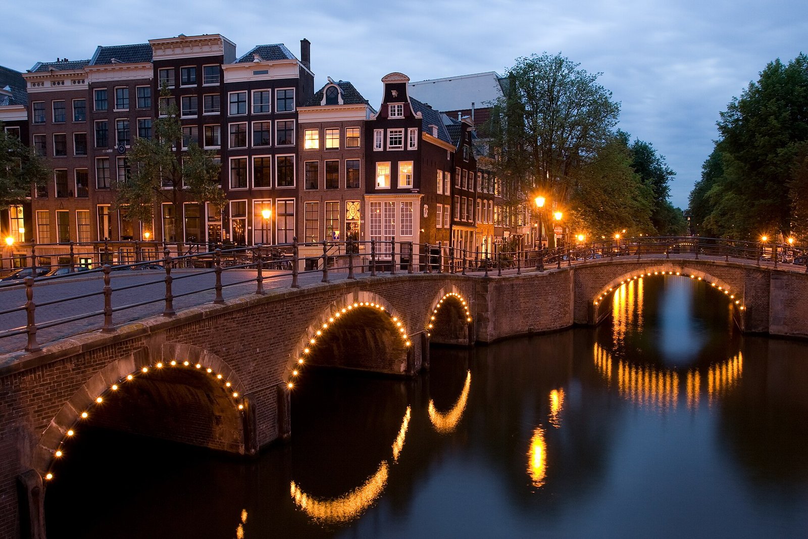 Beleuchtete Backsteinbrücken über die Keizersgracht in der Abenddämmerung, dahinter Grachtenhäuser mit erleuchteten Fenstern