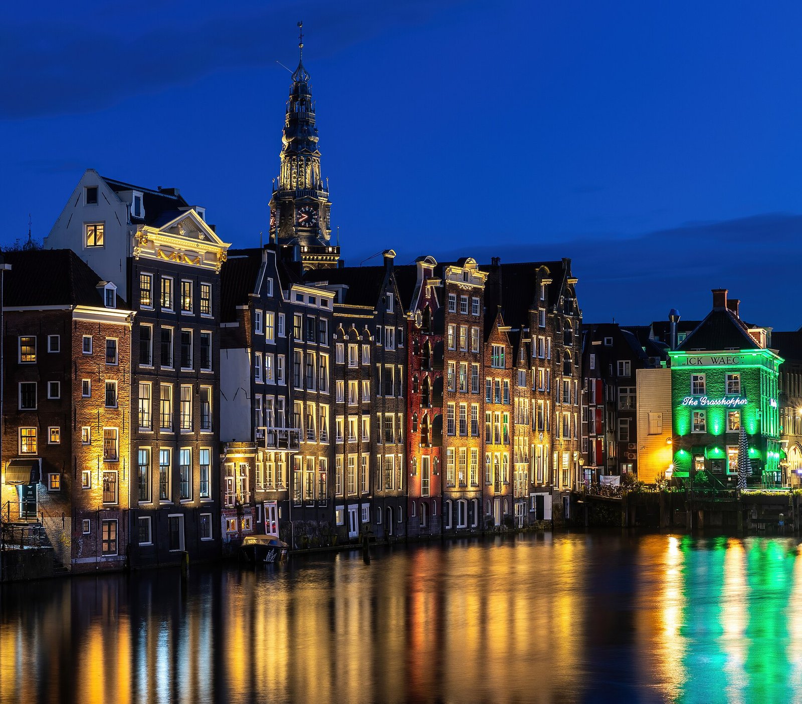 Grachtenhäuser mit erleuchteten Fenstern und ein Kirchturm bei Nacht, grüne Lichter spiegeln sich im Wasser