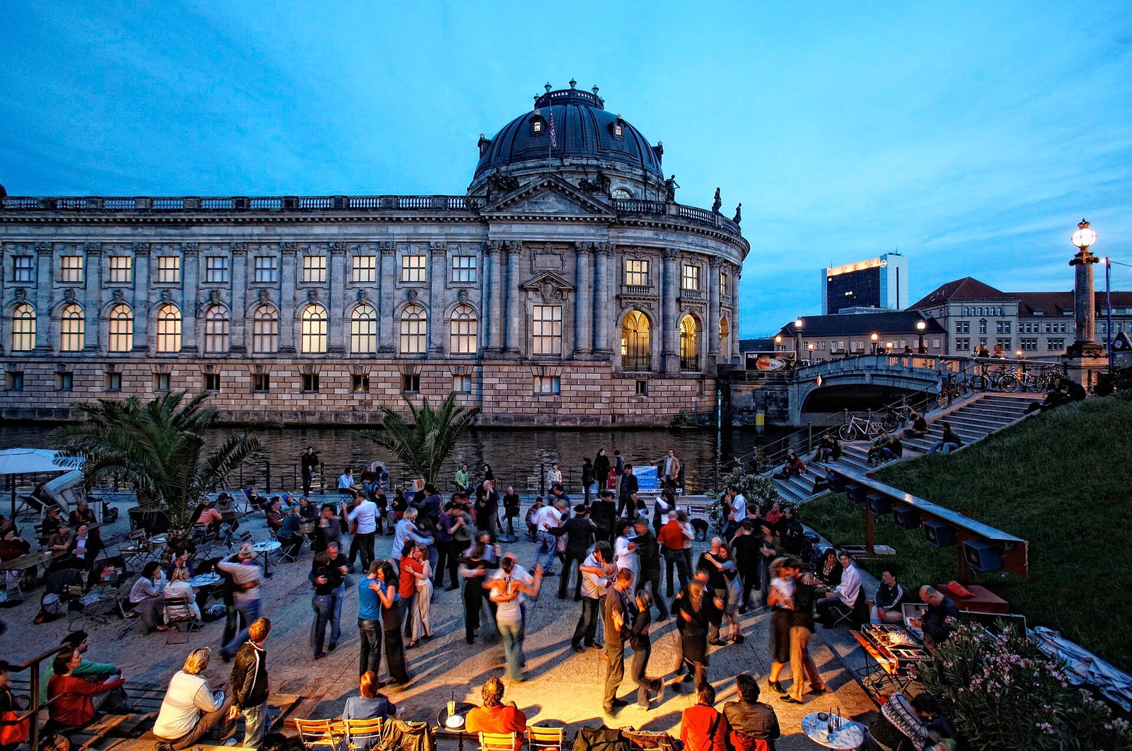 Tanzende Paare auf einer Freifläche an der Spree in der Abenddämmerung, dahinter die Kuppel eines Museums