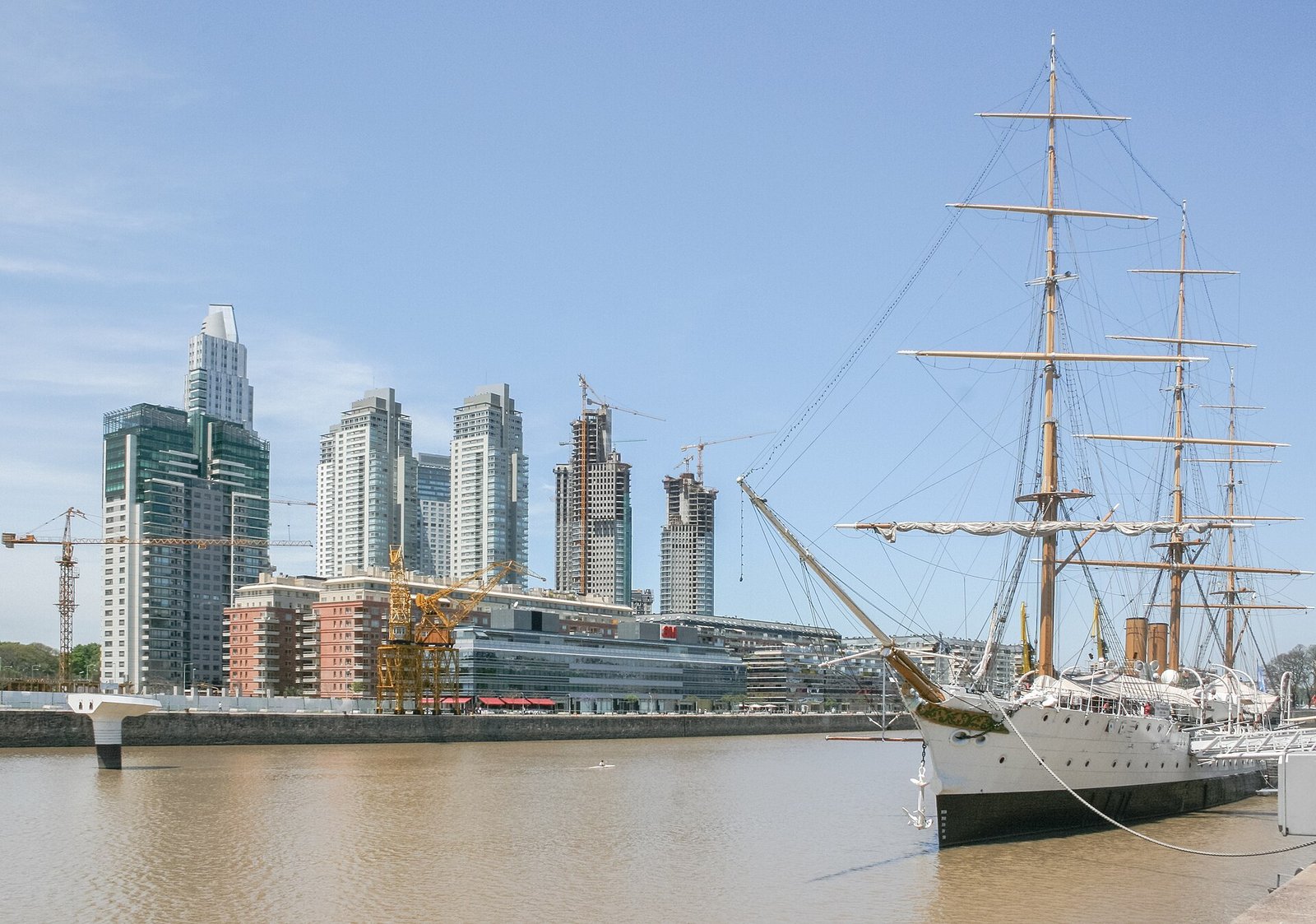 Ein weißes Segelschiff mit hohen Masten am Dock in Puerto Madero, dahinter moderne Hochhäuser und Baukräne