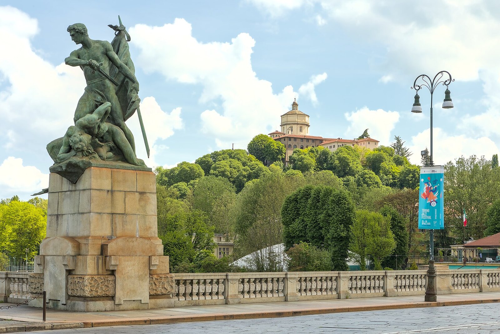 Bronzestatue auf einem Steinsockel an der Brücke Umberto I, dahinter ein grüner Hügel mit einer Kirche