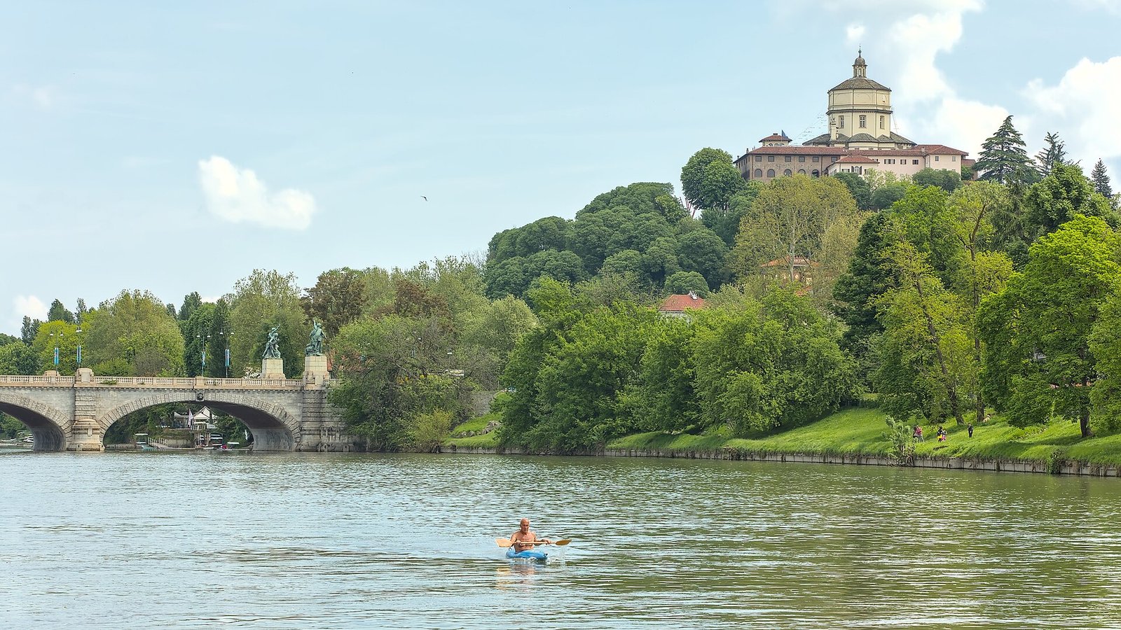 Der Po in Turin mit einer Steinbrücke links und einem bewaldeten Hügel mit einer Kirche auf der Kuppe rechts